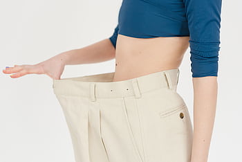 A woman demonstrates significant weight loss by wearing oversized pants indoors. HD wallpaper
