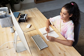 A young girl smiling while studying online at home, using a computer and taking notes. HD wallpaper