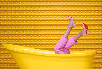 Legs in pink pants and red high heels pop out of a yellow bathtub against a creative rubber duck wall. HD wallpaper