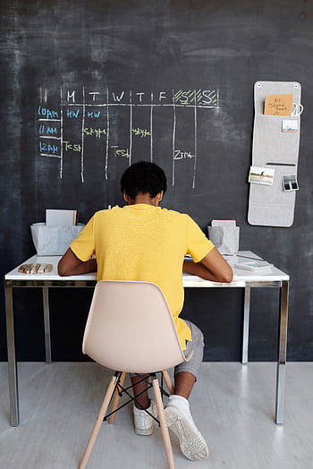 A teenager focuses on studying at a desk, with a blackboard displaying a schedule in the background. HD phone wallpaper