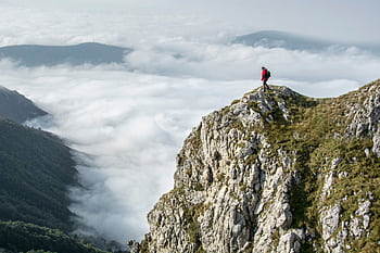 Hiker in red standing on a rocky mountain summit enveloped by clouds. HD wallpaper
