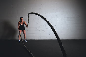 A focused woman performs a dynamic battle rope exercise in a gym setting, demonstrating strength and fitness. HD wallpaper