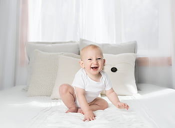 Cute baby sitting and smiling on a white bed in a bright, airy bedroom. HD wallpaper
