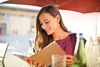 A woman smiles as she reads a menu at an outdoor restaurant, enjoying a sunny day. HD wallpaper