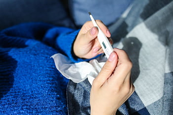 A woman checks a digital thermometer while lying indoors, showing signs of illness. HD wallpaper
