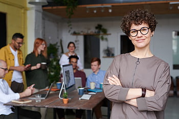 Smiling businesswoman with curly hair stands confidently in a modern office space with colleagues. HD wallpaper