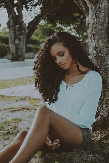 A contemplative woman with curly hair sits outdoors under a tree in a serene park setting. HD phone wallpaper