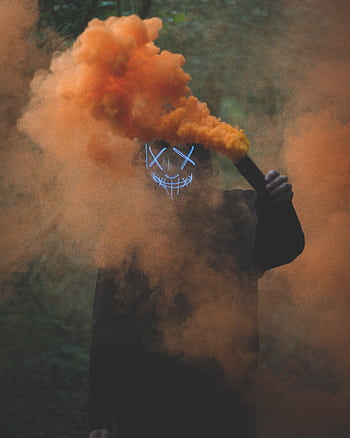 A person wearing an LED mask holds a smoke bomb, enveloped in vivid orange smoke. HD phone wallpaper
