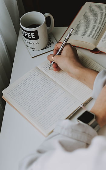 Person writing in a notebook on a desk with a coffee mug, creating a cozy workspace vibe. HD phone wallpaper