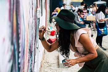 Young woman wearing hat painting a mural on an urban street wall during the day. HD wallpaper