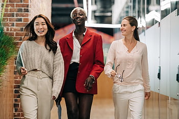 Three diverse and confident businesswomen walking together in a modern office hallway, exuding professionalism and joy. HD wallpaper
