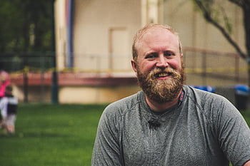A cheerful bearded man smiling outdoors in a casual setting with greenery. HD wallpaper