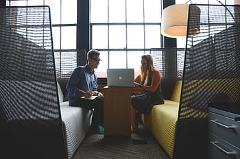 Two adults discussing work and collaborating in a modern office lounge area. HD wallpaper