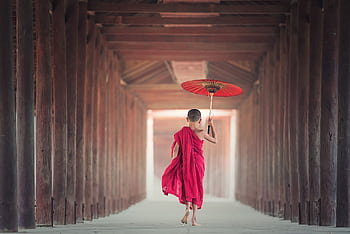 A child monk holding a parasol walks through a traditional Asian temple corridor, embracing cultural heritage. HD wallpaper