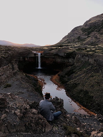 A lone traveler by a waterfall, surrounded by a dramatic landscape. HD phone wallpaper