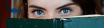 Close-up of a woman with blue eyes peeking over a book, portraying curiosity. HD wallpaper