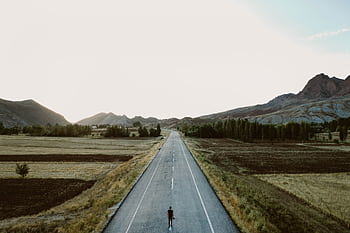 A lone person walks on a rural highway surrounded by fields and mountains under a vast sky. HD wallpaper