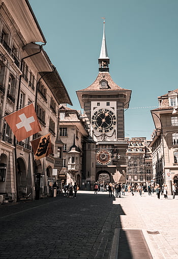 View of Bern's iconic Zytglogge clock tower with Swiss flags and tourists. HD phone wallpaper