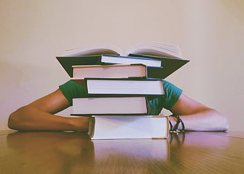 A student hidden behind a stack of books, symbolizing academic stress and study challenges. HD wallpaper