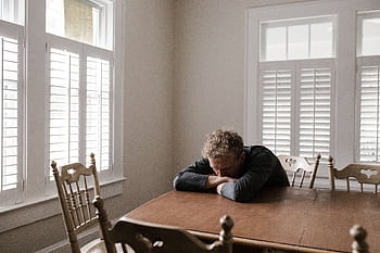 An adult man with emotions of sadness and anxiety sitting alone at a wooden table near windows. HD wallpaper