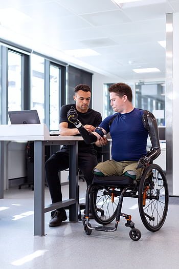 Engineer adjusts prosthetic arm for seated man in wheelchair, focusing on modern technology in healthcare. HD phone wallpaper