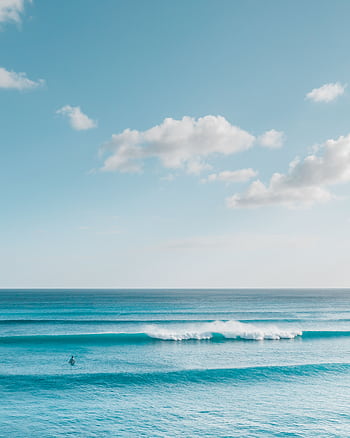 A peaceful ocean scene with gentle waves and a distant surfer under a clear blue sky. HD phone wallpaper