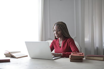 Woman with braided hair smiling as she works on a laptop at home with books around her. HD wallpaper
