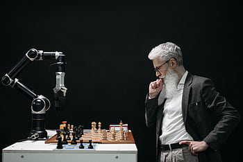 An elderly scientist contemplates a chess move against a robotic arm on a chessboard. HD wallpaper
