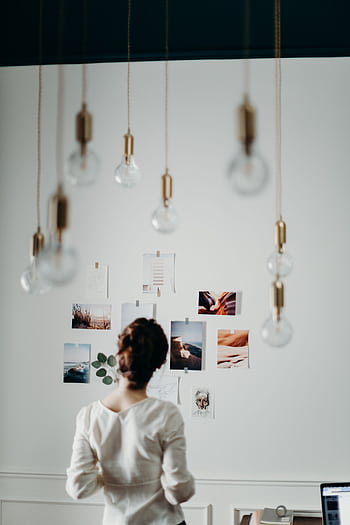 Woman in white top examining mood board under hanging lights in a modern interior design setting. HD phone wallpaper