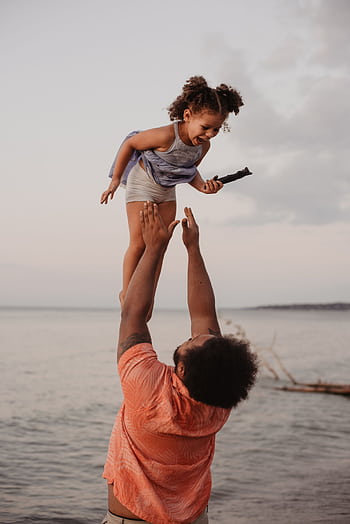 A joyful father lifts his daughter in the air by the beach during sunset, capturing a moment of love and happiness. HD phone wallpaper