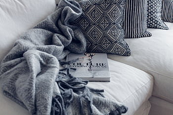 A stylish living room featuring a white sofa with patterned pillows, a blanket, and a fashion book. HD wallpaper