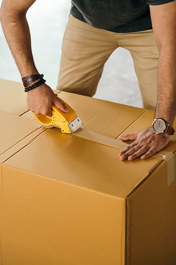 Close-up of a man using a tape dispenser to seal a cardboard box indoors. HD phone wallpaper
