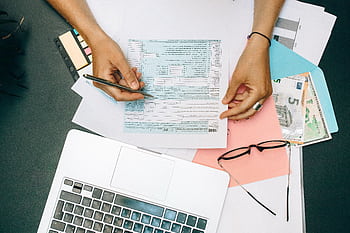 Hands writing on tax documents with laptop, glasses, and currency on desk. HD wallpaper