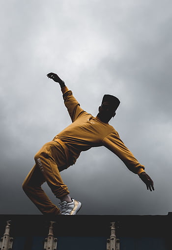 A dancer in vibrant yellow attire executes a dynamic pose against a dramatic cloudy sky, captured in a low angle shot. HD phone wallpaper