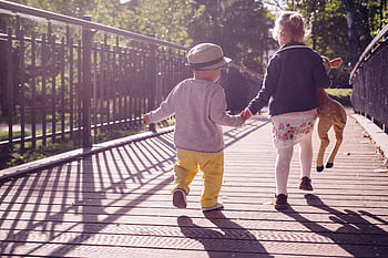 Two children walking hand in hand on a sunlit bridge, holding a stuffed toy deer. HD wallpaper
