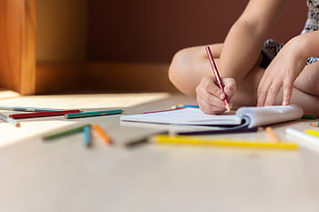 A child sitting indoors, drawing in a notebook with colorful crayons, surrounded by art supplies. HD wallpaper
