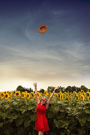 A care woman wearing a red dress and sunglasses joyfully throws her straw hat in a vibrant sunflower field. HD phone wallpaper