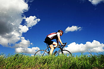 A cyclist in sportswear riding a bike under a bright blue sky with clouds. HD wallpaper