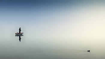 A fisherman stands on a boat in a misty lake, creating a serene reflection. Perfect for peaceful and nature themes. HD wallpaper