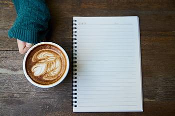 Flat lay of a coffee cup with latte art next to a blank notebook on a wooden table. HD wallpaper