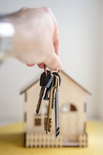 Close-up of a hand holding keys with a miniature wooden house in the background, symbolizing real estate investment. HD phone wallpaper