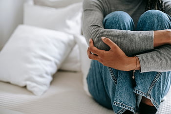 An unrecognizable woman sits on a bed, embracing her knees, conveying solitude and contemplation. HD wallpaper