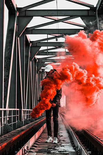 Man holding orange smoke bomb on railway bridge, creating a vibrant and dramatic scene. HD phone wallpaper