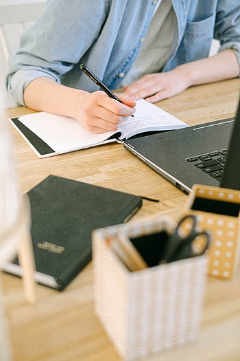 Adult writing notes on a notebook while working on a laptop in a cozy home office environment. HD phone wallpaper