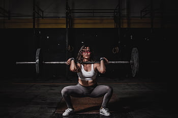 A strong female weightlifter squats with a barbell in a dimly lit gym, showcasing power and fitness. HD wallpaper