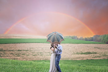 A couple shares a kiss under an umbrella in a scenic rural landscape with a rainbow. HD wallpaper