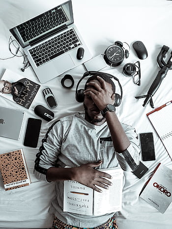 A young man lying on a bed surrounded by modern gadgets, books, and tech devices, illustrating digital lifestyle. HD phone wallpaper