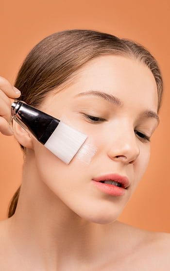 Close-up of a woman applying skincare product with a brush against a neutral background. HD phone wallpaper