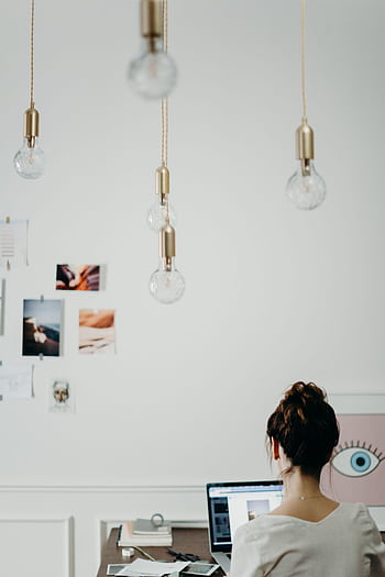Woman working at a desk in a modern creative office, surrounded by mood boards and hanging lightbulbs. HD phone wallpaper