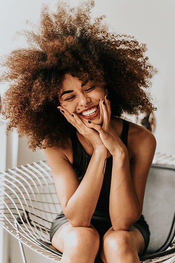 A cheerful woman with an afro hairstyle sitting indoors, showcasing happiness and style. HD phone wallpaper